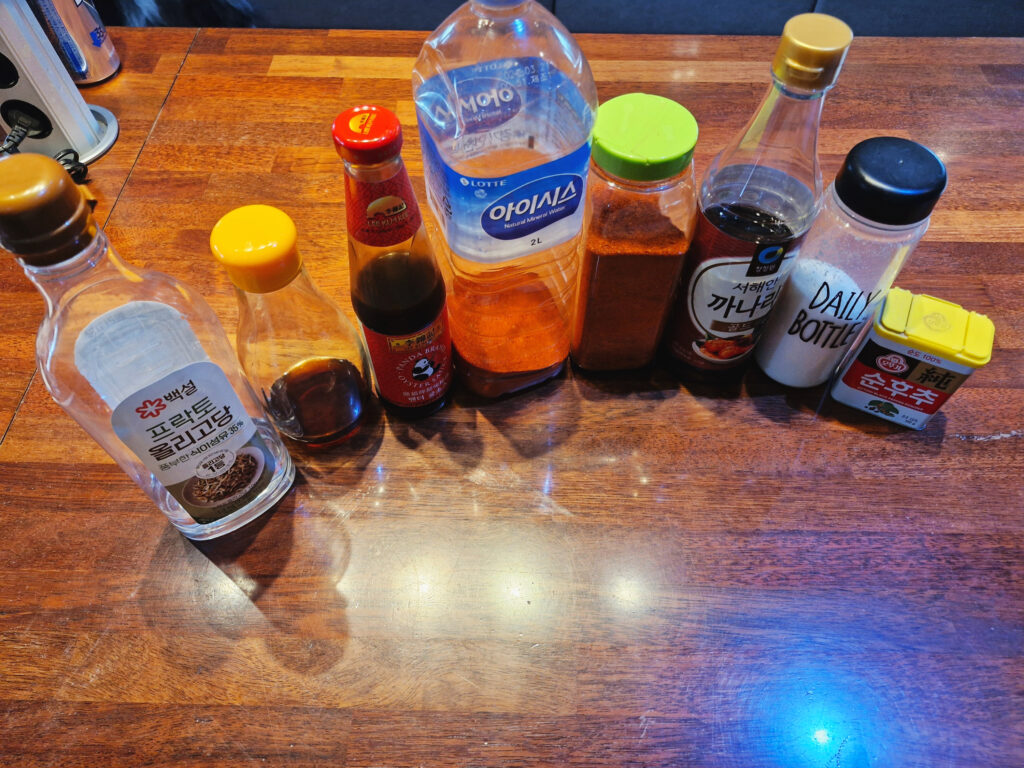 An assortment of authentic Korean marinating ingredients on a wooden table, including Beksul Oligosaccharide, oyster sauce, chili powder (Gochugaru), fish sauce, and various seasonings for the chicken marinade.