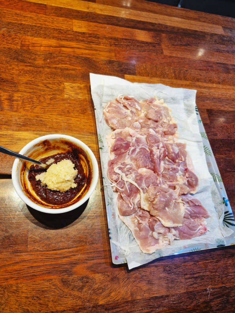 Preparing chicken thighs by drying them with paper towels and mixing the spicy red marinade with minced garlic in a bowl.
