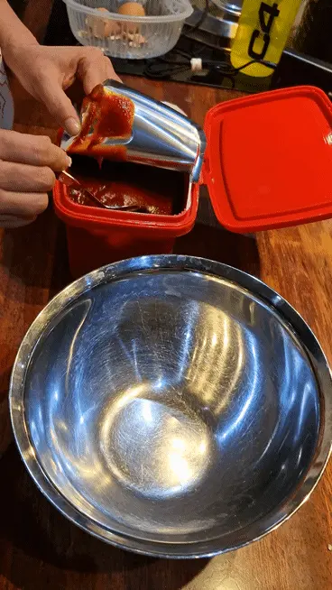 Scoop of Gochujang (Korean chili paste) being added to a stainless steel mixing bowl