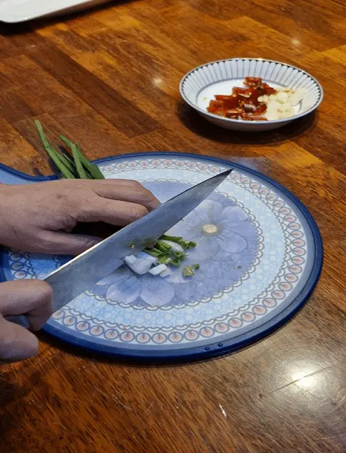 A person thinly slicing fresh garlic and red chilies on a wooden cutting board.